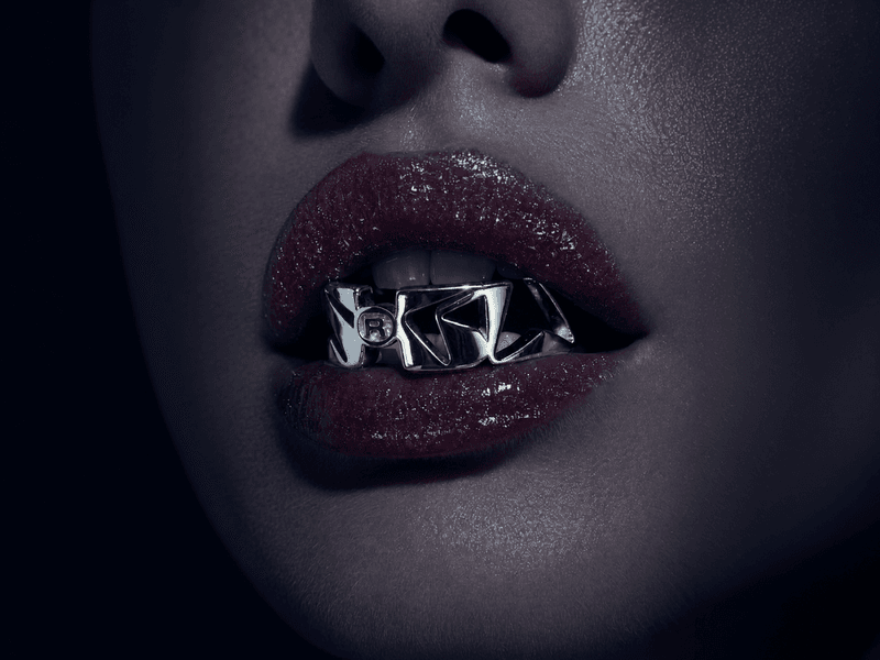Close-up beauty shot of glossy lips wearing a futuristic chrome grill accessory with sculpted metallic lettering in a dark cinematic aesthetic